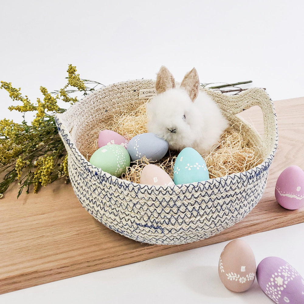 
                      
                        Small white rabbit in a decorative Easter basket with pastel eggs on a wooden surface.
                      
                    