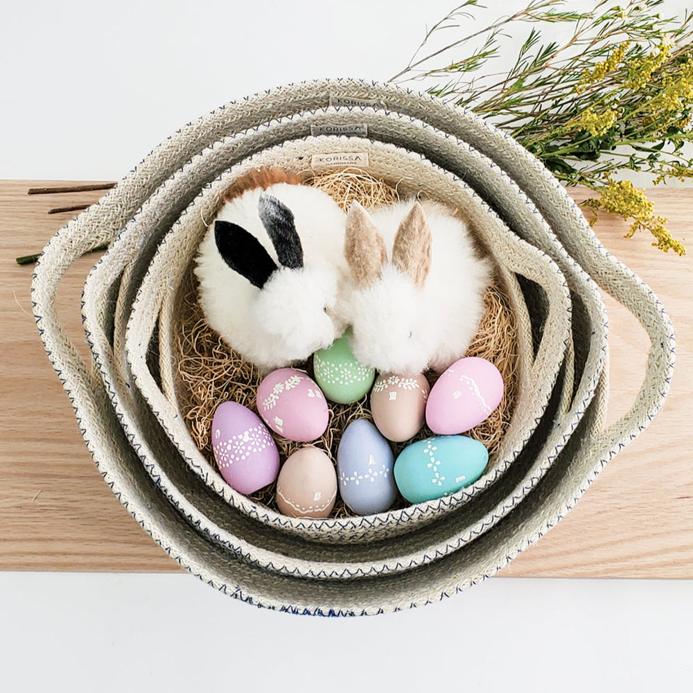 
                      
                        Small rabbit in a nest of pastel eggs inside a woven basket on a wooden surface.
                      
                    