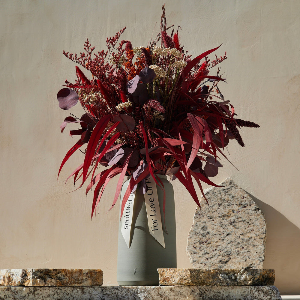 A monochromatic Scarlet Dried Floral Bouquet featuring preserved weeping willow, red limonium, cream rice flowers, and fiery red thistle, displayed in a gray vase on a stone surface.