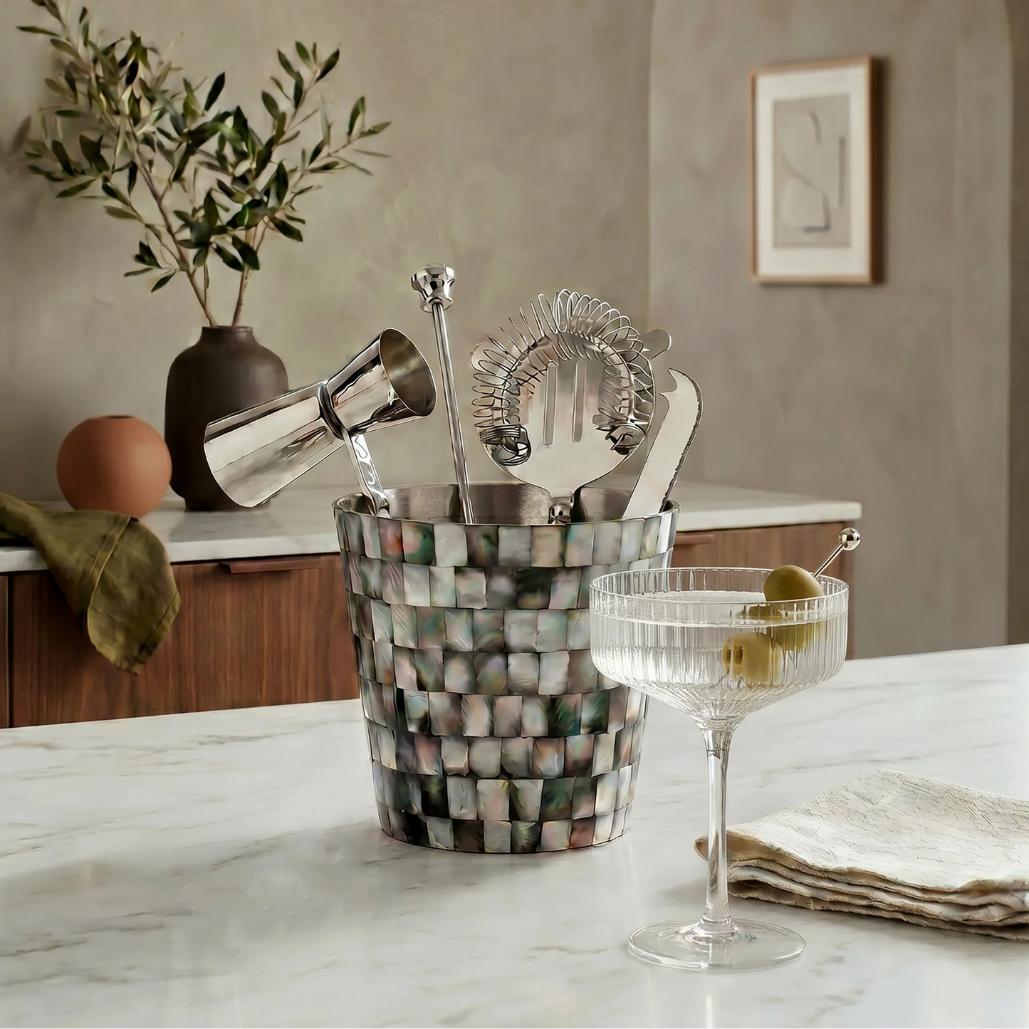 A mottled mosaic cocktail bucket filled with silver bar tools, set on a white marble countertop in a styled kitchen scene with a coupe glass and folded napkin nearby.