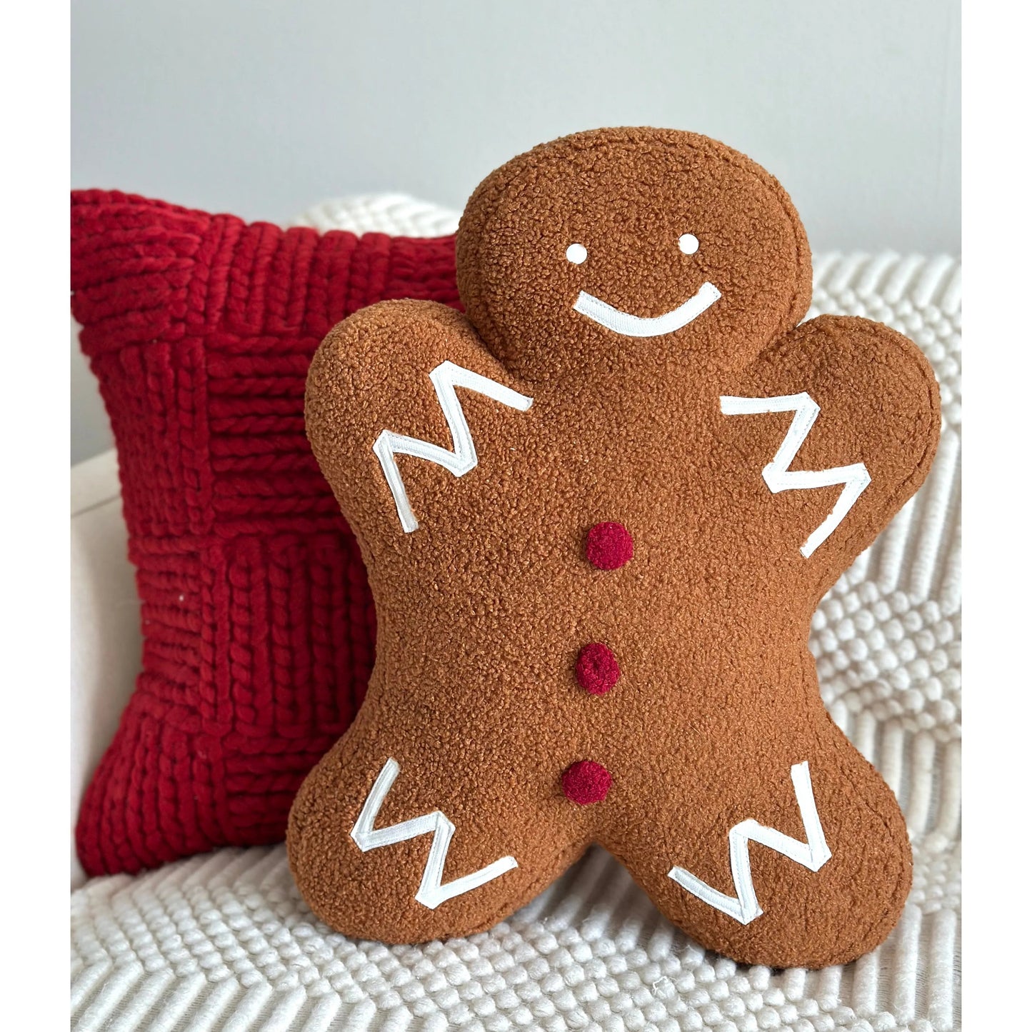 Gingerbread man holiday pillow in brown with white icing details, paired with a red textured cushion, by Busa Designs.