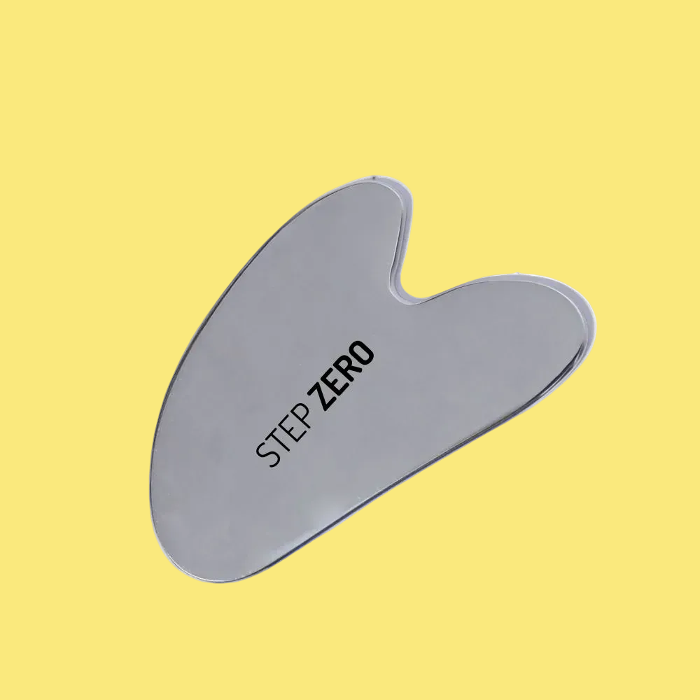 A stainless steel gua sha facial scraping tool with a smooth, polished finish and the text 'STEP ZERO' printed in black on its surface, shown against a plain yellow background.