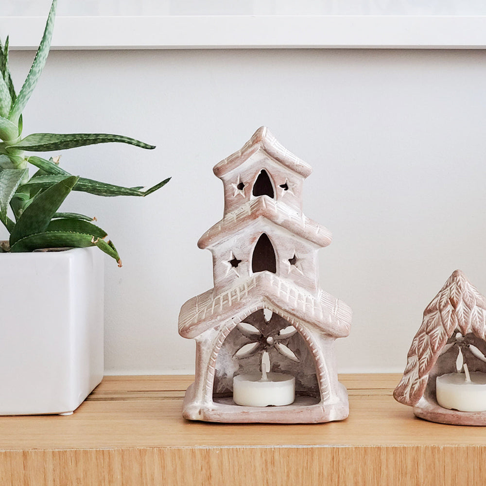 Decorative terracotta tealight candle holder shaped like a house with a candle on a wooden surface next to a plant.