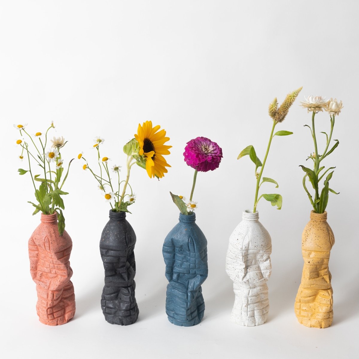 An assortment of concrete water bottle vases in five different colors with flower stems in each.