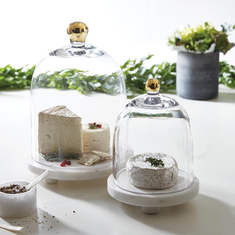 Two glass cloches with gold knobs, large and small, on marble bases covering various desserts