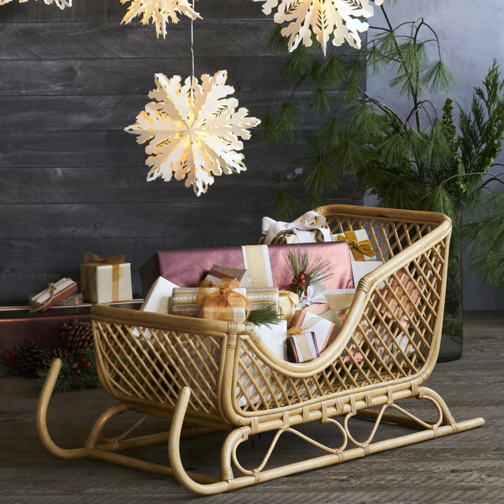 Shelburne™ Woven Rattan Sleigh holding wrapped presents under hanging paper snowflakes